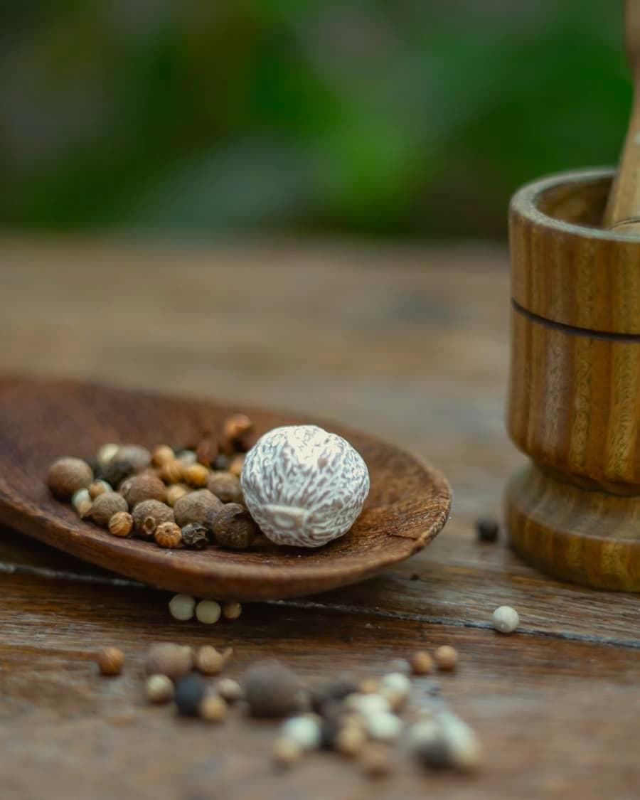 Whole peppercorns and nutmeg on a wooden dish beside a mortar