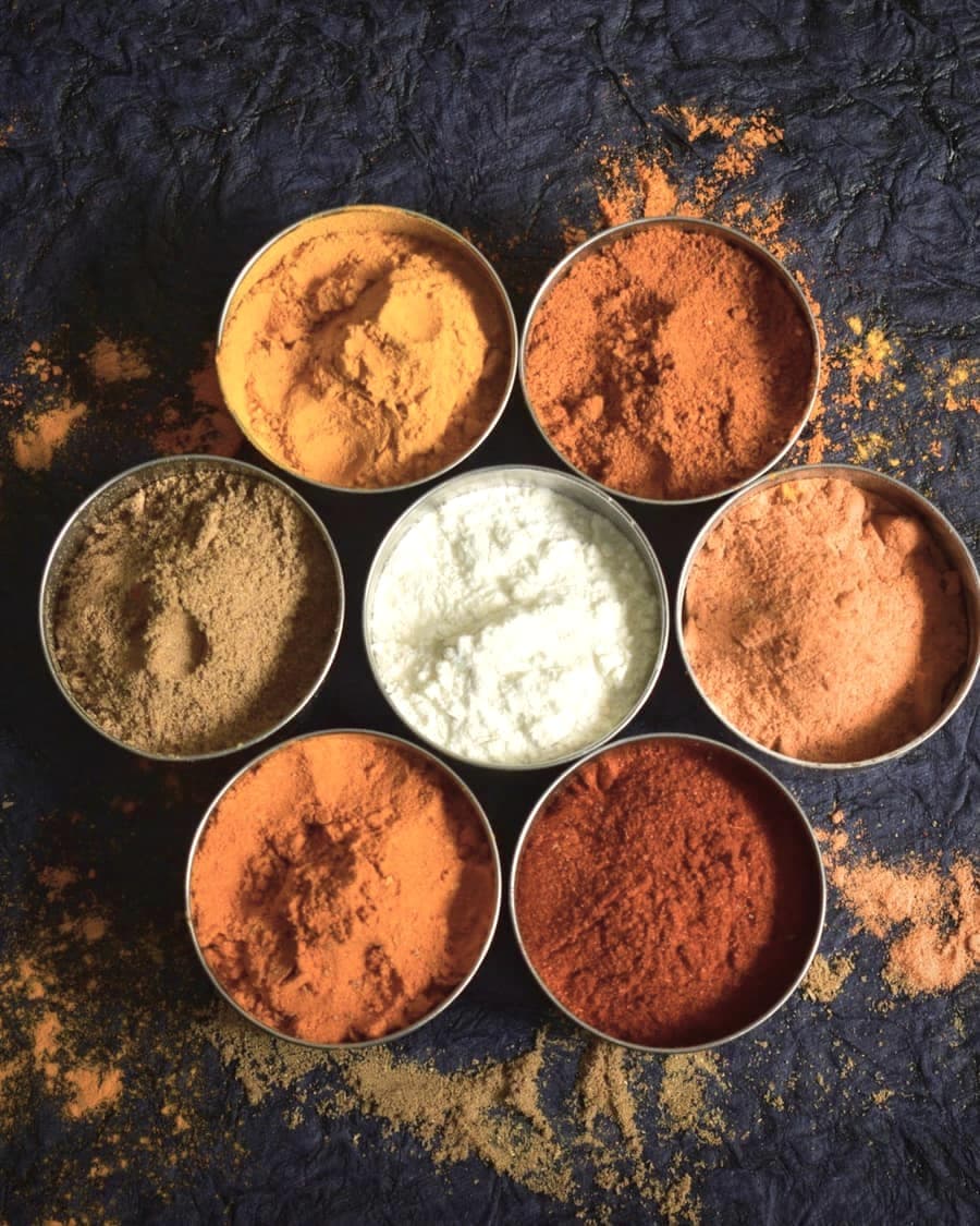 Many small bowls of coloured ground spices on a dark textured surface